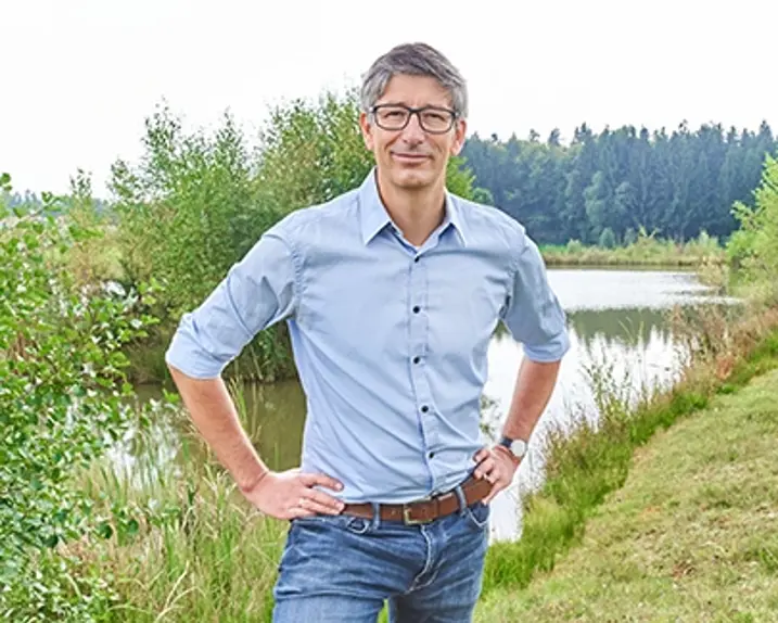 Christian Rucker steht im Grünen vor einem Weiher. Er trägt eine Jeans und ein hellblaues Hemd.