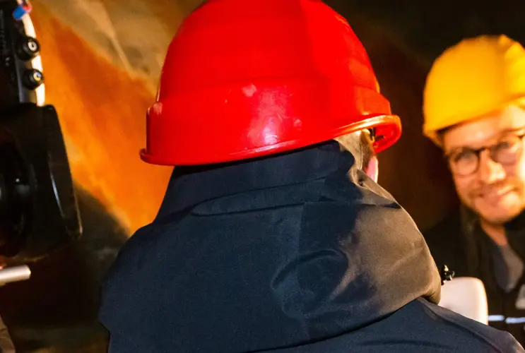 Ein Blick von Hinten auf eine Interviewsituation. Ein Mann mit einem roten Schutzhelm interviert einen Mann mit Brille und gelben Schutzhelm. Links im Bild sieht man eine Kamera.